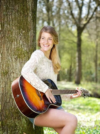 The girl rests the guitar on her thigh while leaning against the tree.